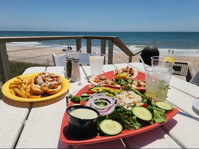 Image from Instagram @daddymacsbeachgrille A plate of seafood salad and a shrimp po'boy sandwich with fries on an occeanside table in surf city